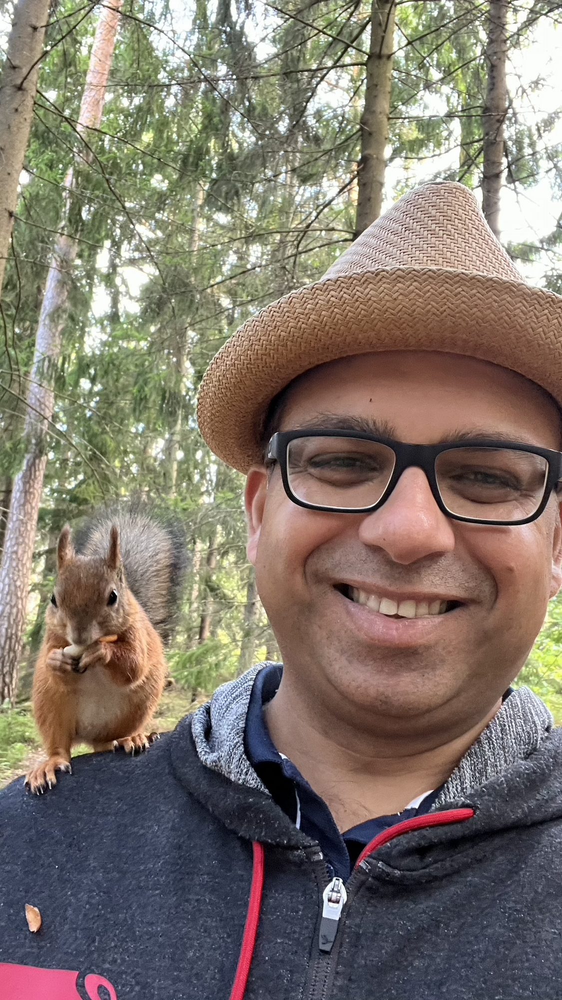 Anupam smiling in a straw hat while a red squirrel sits on his shoulder eating a nut, in a pine forest.